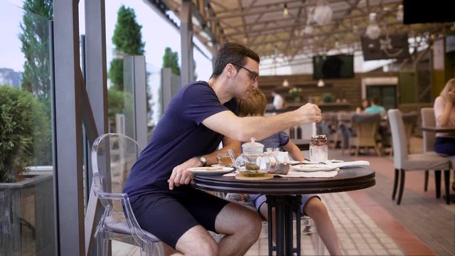 Handsome man in casual blue shirt and breeches sitting in restaurant with son and people in the background. Young father eating dessert of child with long teaspoon. Little boy talking to male parent.
