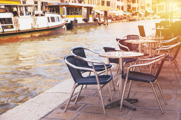 Fototapeta na wymiar Restaurant tables and chairs with vaporetto ships in the background in Venice, Italy. European travel, outdoor dining and cuisine.
