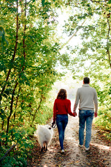 a young couple with a dog walking along the autumn park she is in a red sweater he is in white with a husky samoyed dog, a white dog and love