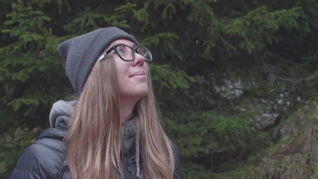 Young blonde girl wearing glasses in wonder at the snowflakes falling around her outdoors in the pine forest. Slow motion shot of attractive woman enjoying first snow in a park.