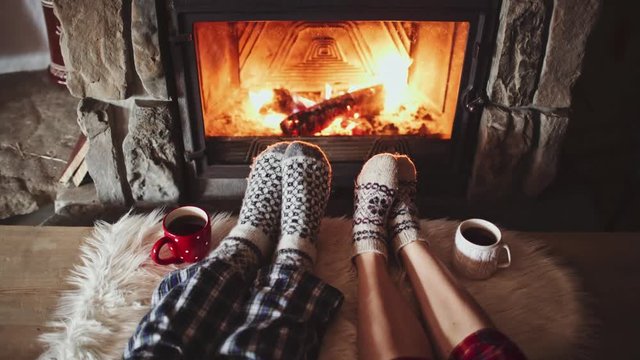 Couple Feet in Woollen Socks by the Cozy Fireplace, 4K. Man and Woman relax by warm fire and warming up their feet. Close up. Winter and Christmas holidays concept. 