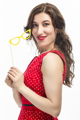 Sensual Alluring Caucasian Female Holding Yellow  Artistic Spectacles In Front of Cheek. Posing  In Red Polka Dot Dress Against Pure White.