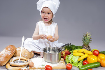 Surprised Caucasian Little Girl In Cook Uniform With Fresh Eggs and Saucepan In Studio with Vegetables At Background. Posing with Angry Expression. Against Gray.