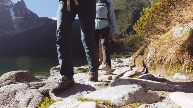 Family Hiking in High Mountains on Cold Sunny Morning. STABILIZED 4K SHOT. Three adult tourists with backpacks walking on a rocky trail by lake with snow-capped mountains. Spring, autumn or winter.  