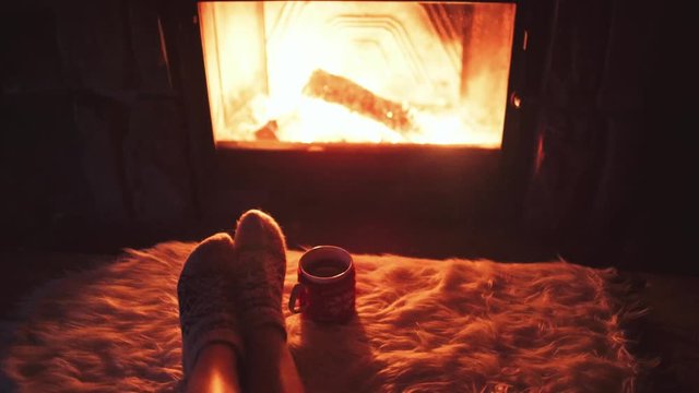 Feet in woollen socks by Burning Christmas Fireplace. 4K STABILIZED shot. Woman relaxes by warm fire with a cup of hot drink and warming up her feet. Close up. Winter and Christmas holidays concept.