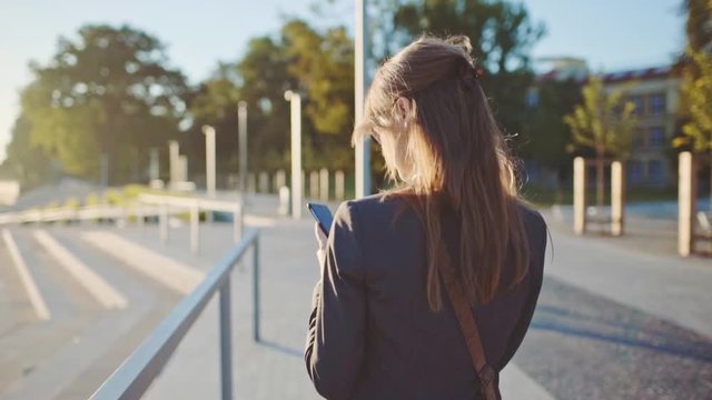 Young Businesswoman Using SmartPhone, Going to Work in the Sunny Morning City. SLOW MOTION. STEADICAM Stabilized Shot. Attractive Professional Business Woman with a cell phone in a city. Lens Flare. 