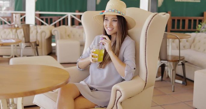 Beautiful elegant woman wearing stylish straw hat and dress holding glass with refreshing drink while relaxing in armchair in resort area.