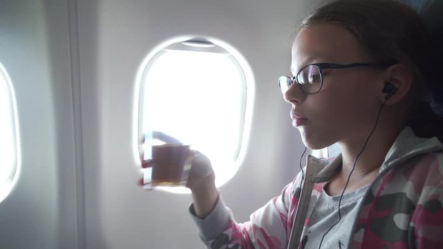 Young girl with glasses and headphones watches video on the monitor built into armchair and drinking juice in the cabin of the airplane stock footage video