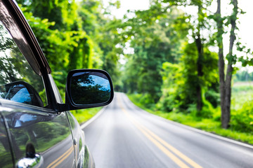 Rear view mirror of gray color car on outback road in the morning time with sunlight and blurred green forest in the background