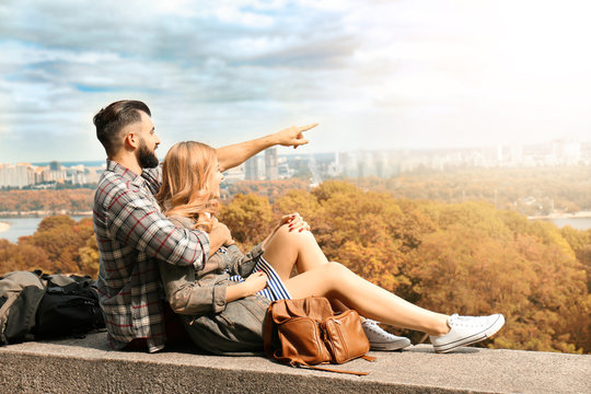 Young couple sitting on parapet in park and enjoying view of beautiful cityscape