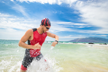 Smartwatch triathlon swimming sport man finishing swim checking heart rate on smart watch. Male triathlete swimmer running out of ocean. Professional athlete in triathlon suit training for ironman.
