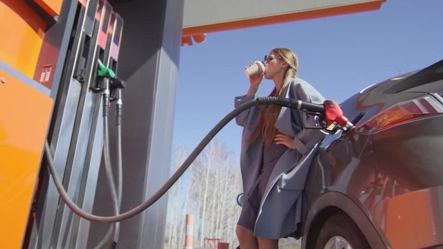 Stylish young lady in sunglasses drinking coffee from disposal cup and looking at gas pump while filling up car at sunny day