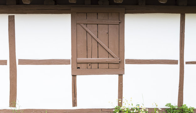 Detail view of a frame house with half-timbered white wall, light brown wooden beams and closed window shutter. Marguerites are growing on the side.