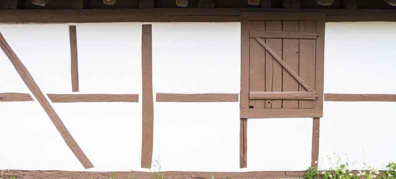 Detail view of a frame house with half-timbered white wall, light brown wooden beams and closed window shutter. Marguerites are growing on the side.
