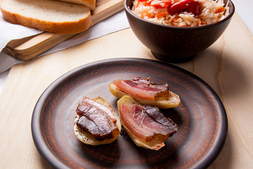 Close up view of baked potatoes with slices of bacon on wooden background. Sauerkraut in clay bowl and slices of white bread on cutting board.