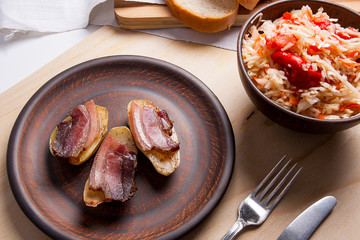 Baked potatoes with slices of bacon and eating utensils on wooden background. Sauerkraut in clay bowl and slices of white bread on cutting board..