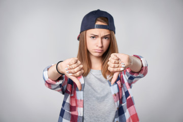 Closeup of discontent teen girl in checkered shirt and baseball cap showing double dislike gesture, over grey background. Shallow depth of field, focus on gesture