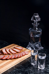 Bottle and shot glasses with vodka, slices of smoked meat and smoked sausage on wooden cutting board.