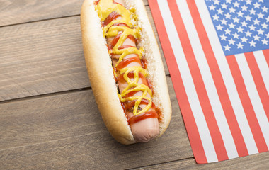 Hotdog with ketchup and mustard next to American flag on wooden table. Fastfood.