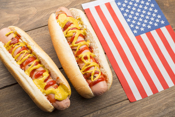 Two hotdogs with ketchup and mustard next to American flag on wooden table. Fastfood.