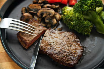 beef steak is cut with knife and fork, with vegetables like broccoli, mushrooms and tomatoes, low carb diet dinner on a dark gray plate