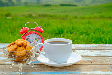 White  coffee cup and cookies  with red alarm clock on wooden table with nature background