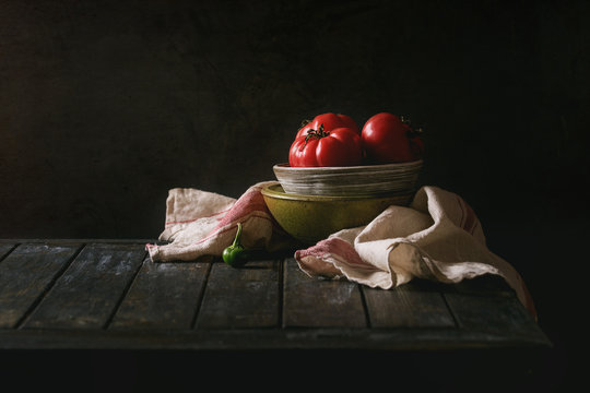 Bowl of tomatoes with pepper and linen towel on old wooden kitchen table. Dark rustic still life.