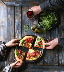 Hands taking sliced homemade pizza with cheese and bresaola, served on black plate with fresh arugula, olive oil, glass of red wine and kitchen towel over old wooden plank background. Flat lay.