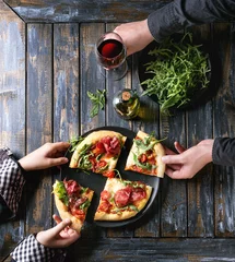 Poster de jardin Pizzeria Hands taking sliced homemade pizza with cheese and bresaola, served on black plate with fresh arugula, olive oil, glass of red wine and kitchen towel over old wooden plank background. Flat lay.