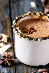 Vintage mug of hot chocolate, decor with nuts, caramel, spices. Ingredients above. Chopped dark and white chocolate, cocoa beans, anise over old wooden table. Dark rustic style. Close up