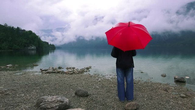 Woman with red umbrella contemplates on rain in front of a lake. Sad and lonely female person looking into distance.