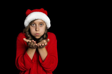 A young girl in a red sweater and a Santa Claus hat blows on her hands and looks at the viewer