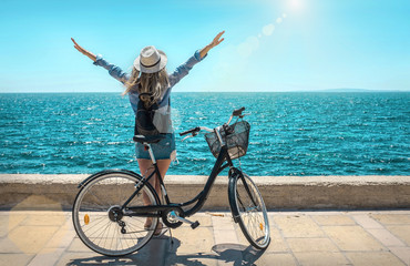 Blonde woman in summer hat with her bicycle walking coastline by