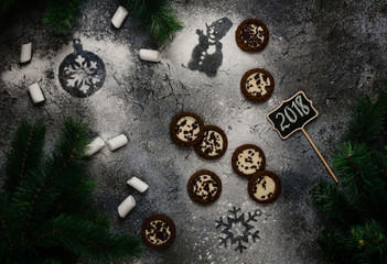 cookies and marshmallows with snowman and snowflake prints on dark blue background with 2018 writing on wooden sign among pine branches, top view