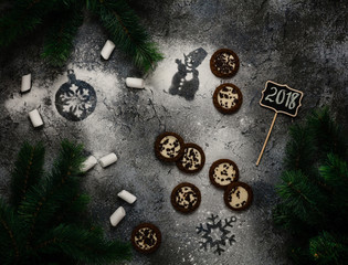 cookies and marshmallows with snowman and snowflake prints on dark blue background with 2018 writing on wooden sign among pine branches, top view