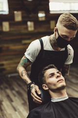 Crop stylist preparing young tattooed man in chair for shaving in barbershop.