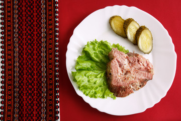 piece of meat with slices of salted cucumber on white plate on the background of a red tablecloth with embroidered napkin