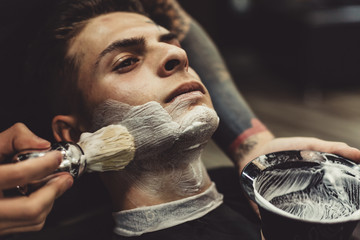 Crop stylish applying foam on customer's cheeks for shaving while working in barbershop.