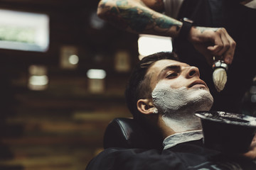 Crop stylish applying foam on customer's cheeks for shaving while working in barbershop. - obrazy, fototapety, plakaty