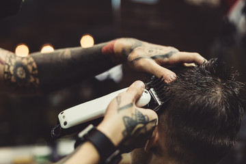 Crop tattooed stylist concentrated on shaving man with machine doing hairstyle.