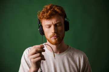 Close-up of young curly readhead hipster holding cord of headphones, with closed  eyes