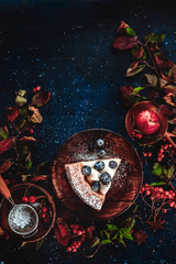 A piece of pumpkin pie with blueberries and persimmon on a dark wooden background with floral decoration. Autumn flat lay with copy space. Conceptual stylised table top shot on dark background.