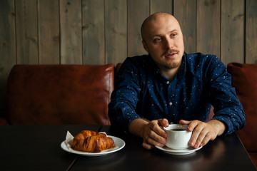 Young serious fashionable man sitting alone in loft-styled cafe and talking with somebody. Cup of coffee and croissants on the table. Former factory building, natural daylight.