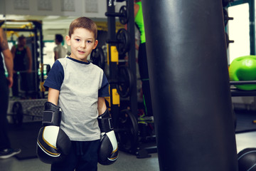 Boy with boxing gloves in the gym near the punching bag. Boy and boxing bag on gym background. Winner fight. Champion...