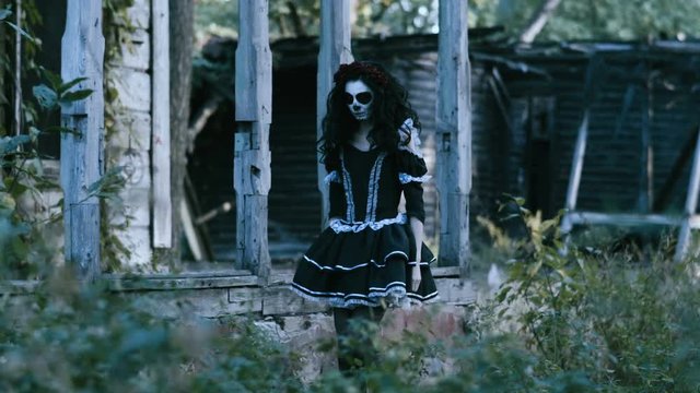 The Mexican Day of the Dead. The young woman with scary make-up of skeleton for Halloween dressed in black clothes going along the wooden abandoned house. 4K