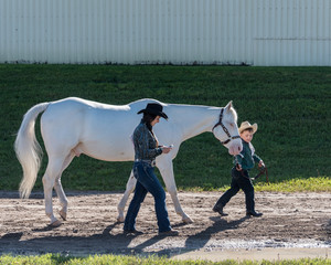 Cowgirl on cellphone as child cowboy leads white appaloosa horse