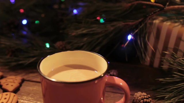 Delicious Christmas holiday with cup of latte. Just made hot drink stays on wooden table on festive background of illuminated pine, gift box and gingerbread scones nearby, close up