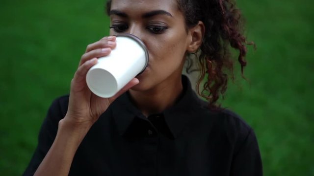 footage of pretty curly african-american woman in black dress with makeup enjoying coffee while working in public park using laptop in slow motion