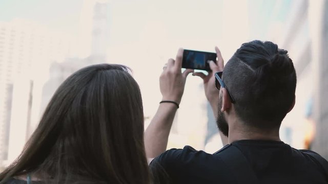 Young beautiful couple walking in downtown of Chicago, America and taking photo of skyscraper on smartphone.