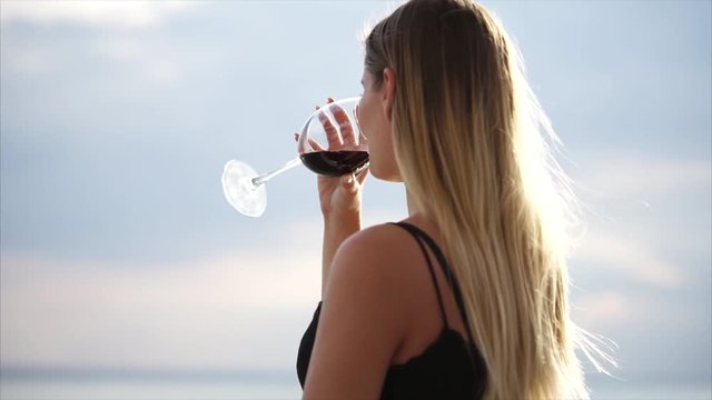 A young and pretty woman drinks red wine from a glass goblet, maybe a lady is in a restaurant with an open terrace, she enjoys nature